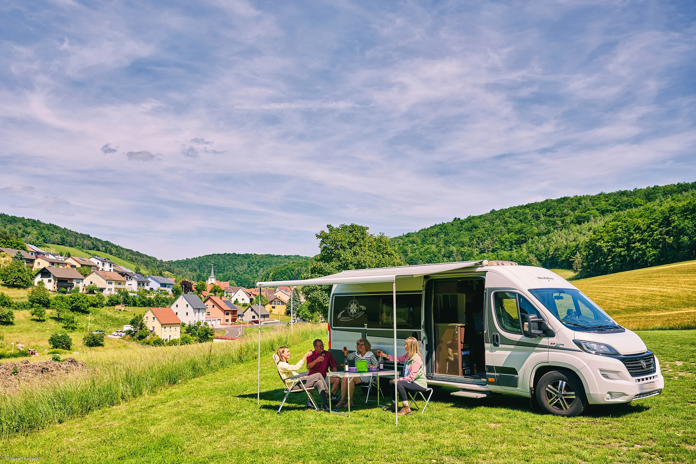 Vier Personen sitzen an einem Tisch unter einer Markise neben einem weißen Wohnmobil auf einer Wiese bei Dorf und Wald