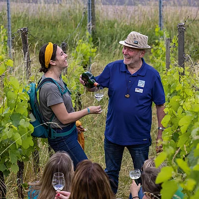 Mann schenkt Frau in Weinberg Weißwein in Glas ein, mehrere Personen beobachten die Szene.