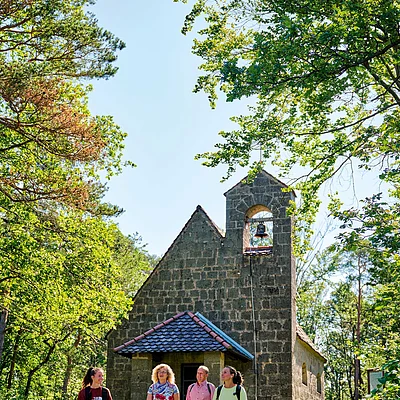 Vier Wanderer gehen auf einem Waldweg vor einer kleinen Steinkapelle mit Glockenturm bei Sonnenschein.
