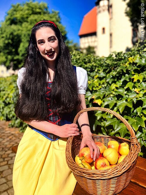 Frau in Schneewittchen-Kostüm hält Korb mit Äpfeln, im Hintergrund grüne Hecke und Gebäude.