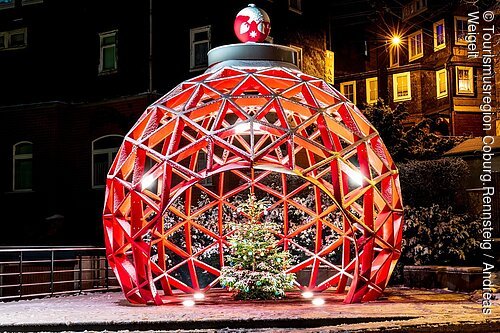 Große rote Weihnachtskugel aus Metall mit beleuchtetem Weihnachtsbaum darin, Schnee und Häuser im Hintergrund.