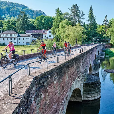 Fünf Radfahrer fahren auf einer alten Steinbrücke über einen Fluss, umgeben von Bäumen und Häusern.
