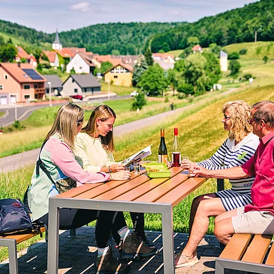 Vier Personen sitzen an einem Tisch im Freien mit Blick auf ein Dorf und grüne Hügel im Hintergrund.