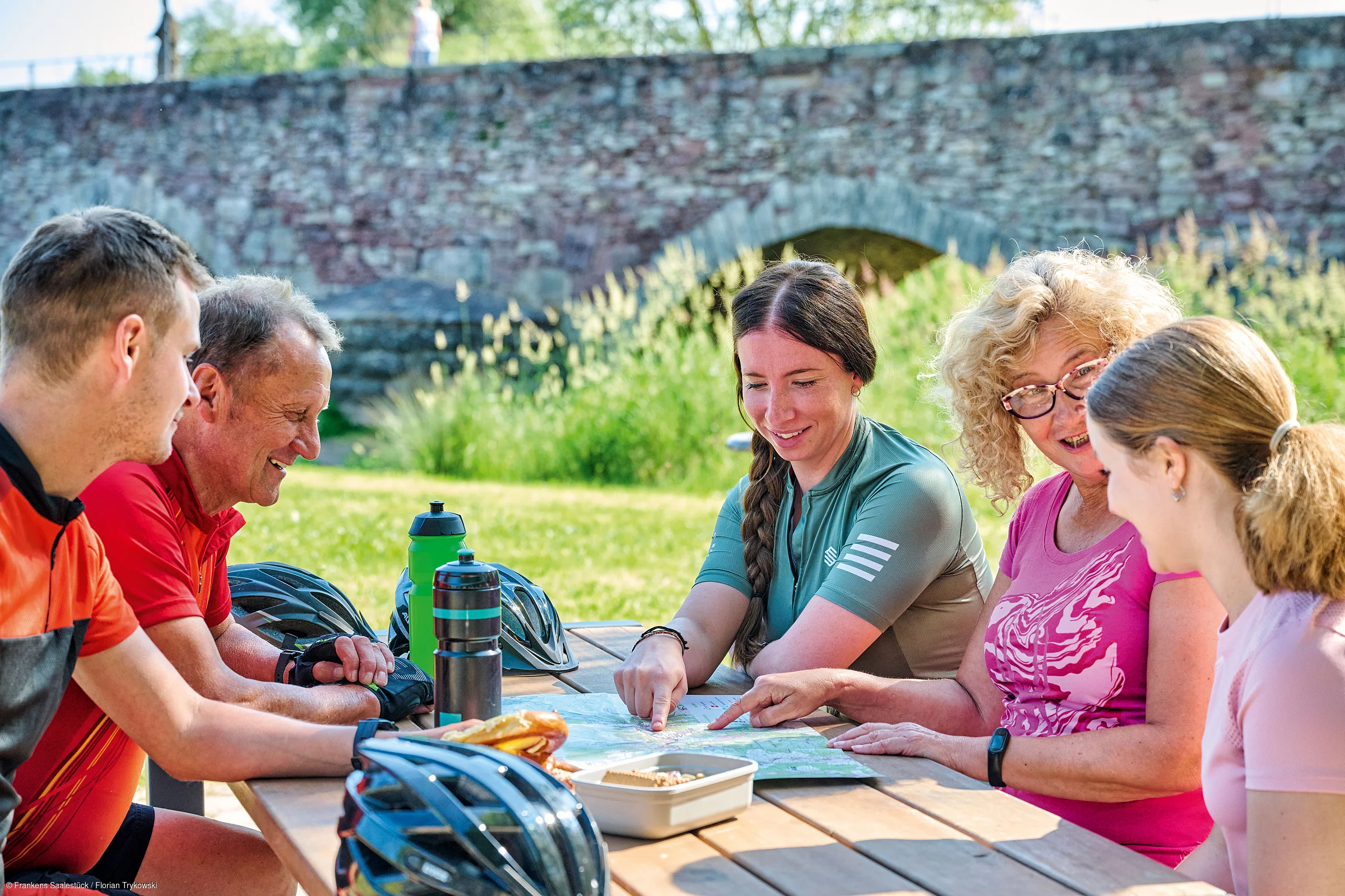 Fünf Personen sitzen an einem Holztisch im Freien und schauen gemeinsam auf eine Karte.