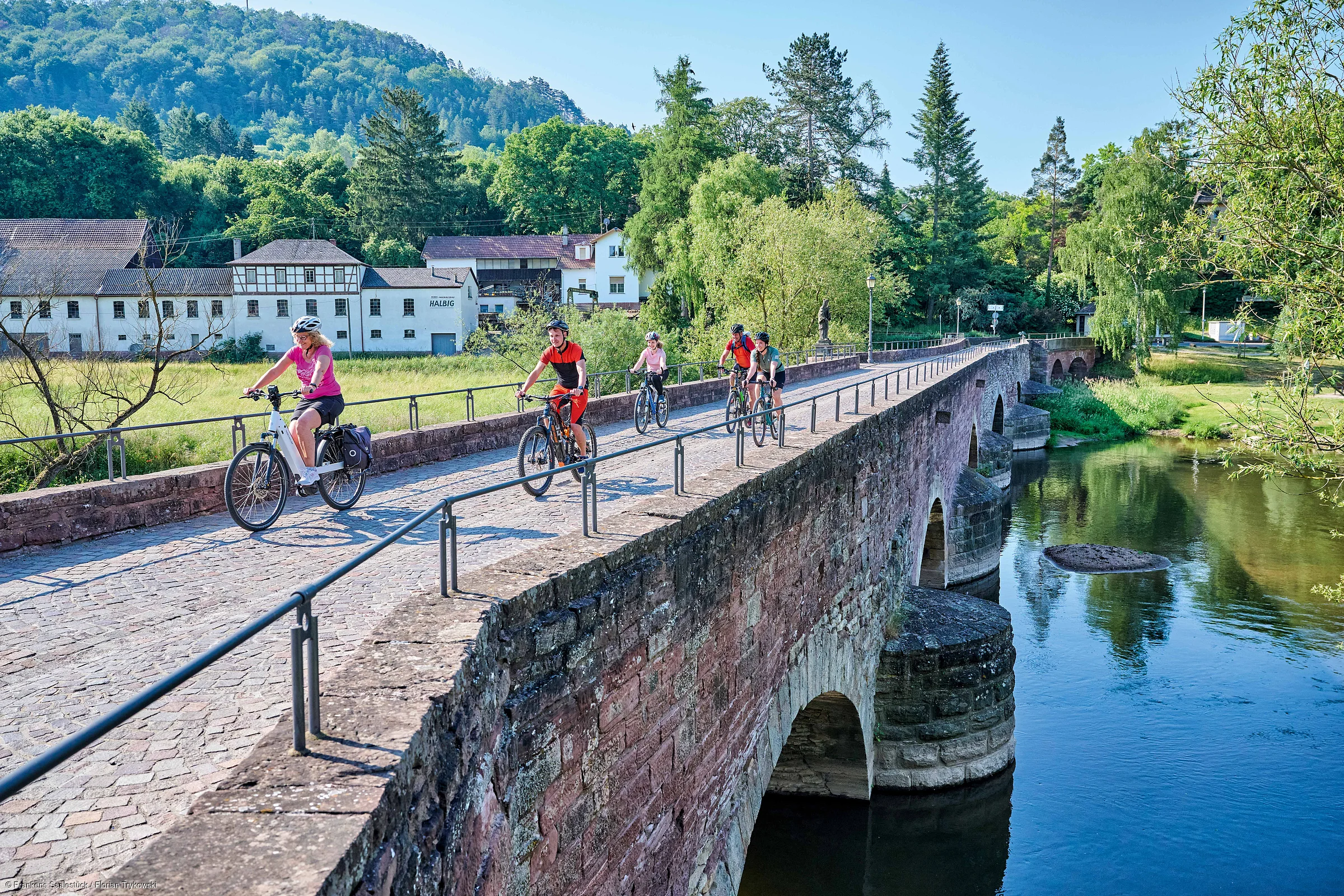 Fünf Radfahrer fahren auf einer alten Steinbrücke über einen Fluss, umgeben von Bäumen und Häusern.