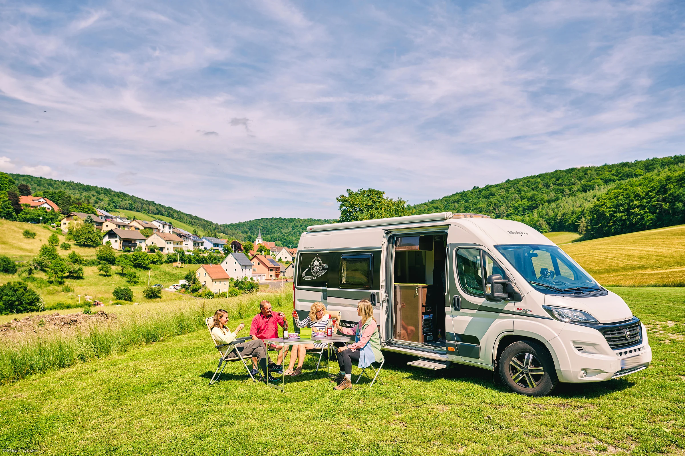Familie sitzt an Campingtisch neben offenem Wohnmobil auf Wiese mit Dorf und Hügeln im Hintergrund.