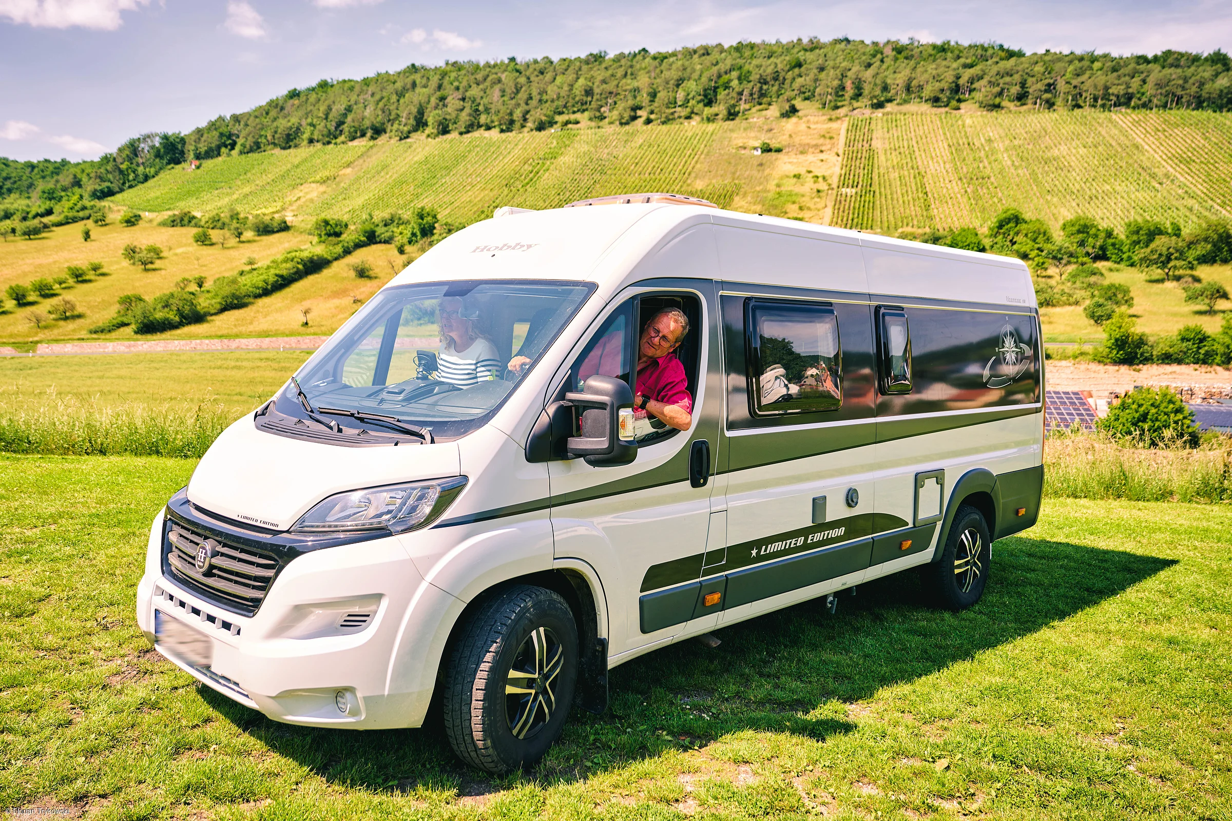 Weißer Wohnmobil-Van mit zwei Personen auf einer grünen Wiese vor bewaldeten Hügeln und Weinbergen.