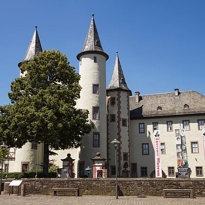 Burg mit drei runden Türmen und Bannern des Weserart-Museums bei klarem Himmel und Bäumen davor.
