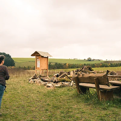 Frau mit Hund auf Wiese vor Holzbankgruppe und Infotafel in ländlicher Umgebung bei bewölktem Himmel.