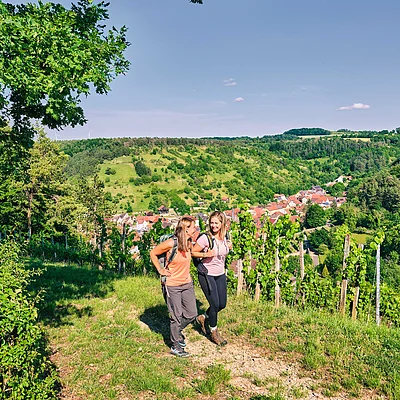 Zwei Frauen mit Rucksäcken wandern auf einem Weg mit Blick auf ein Dorf und bewaldete Hügel im Hintergrund.