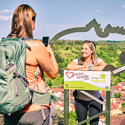Frau mit Rucksack fotografiert eine andere Frau, die hinter einem Rahmen mit Text in ländlicher Umgebung posiert.