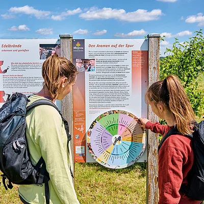 Zwei Frauen mit Rucksäcken lesen eine Infotafel über Edelbrände in einer ländlichen Umgebung bei Sonnenschein.