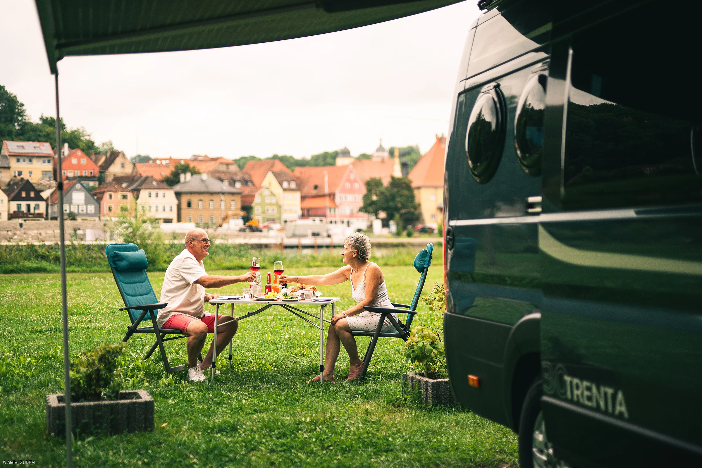 Älteres Paar sitzt an Campingtisch auf Wiese und stößt mit Weingläsern vor einem Wohnmobil an.