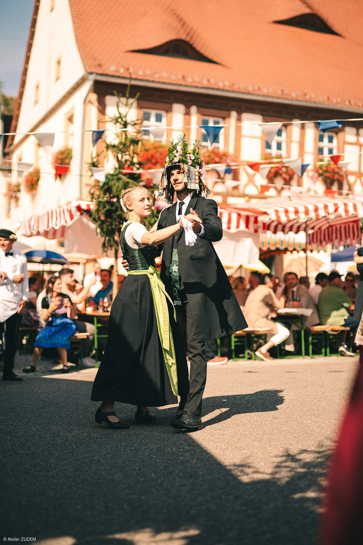 Paar in traditioneller Tracht tanzt auf einem Fest vor einem Fachwerkhaus mit bunten Fahnen und Marktständen.