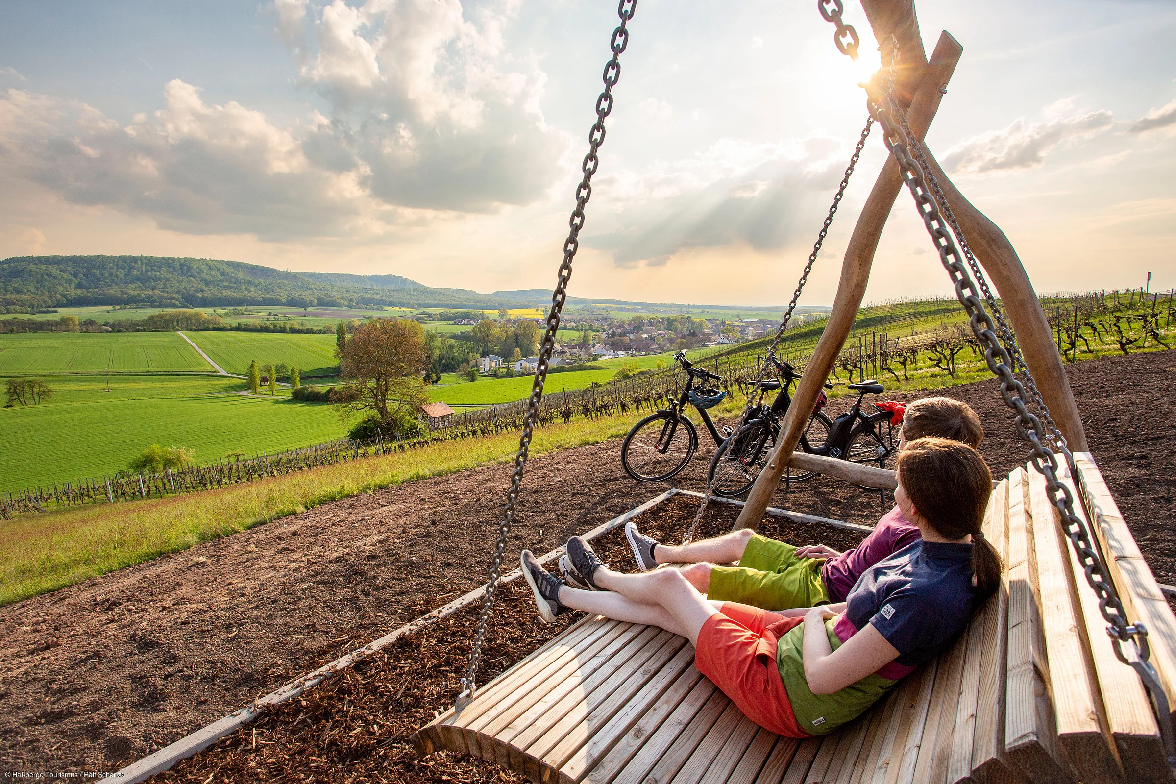 Zwei Personen sitzen auf einer Holzschaukel mit Blick auf grüne Felder und einen Ort im Hintergrund bei Sonnenlicht.