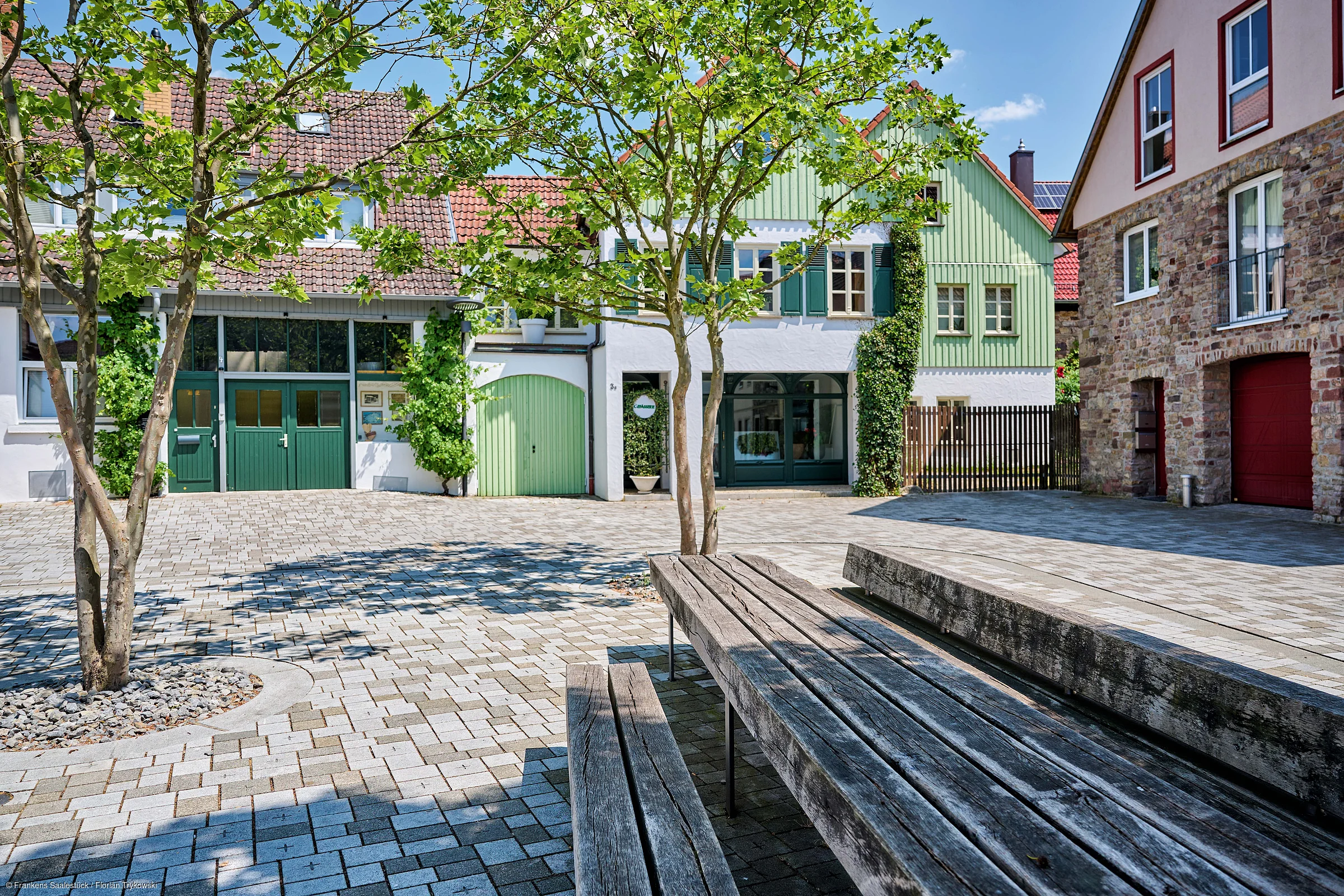 Platz mit gepflastertem Boden, Bänken, Bäumen und Häusern mit grünen Türen und Fensterläden bei Sonnenschein