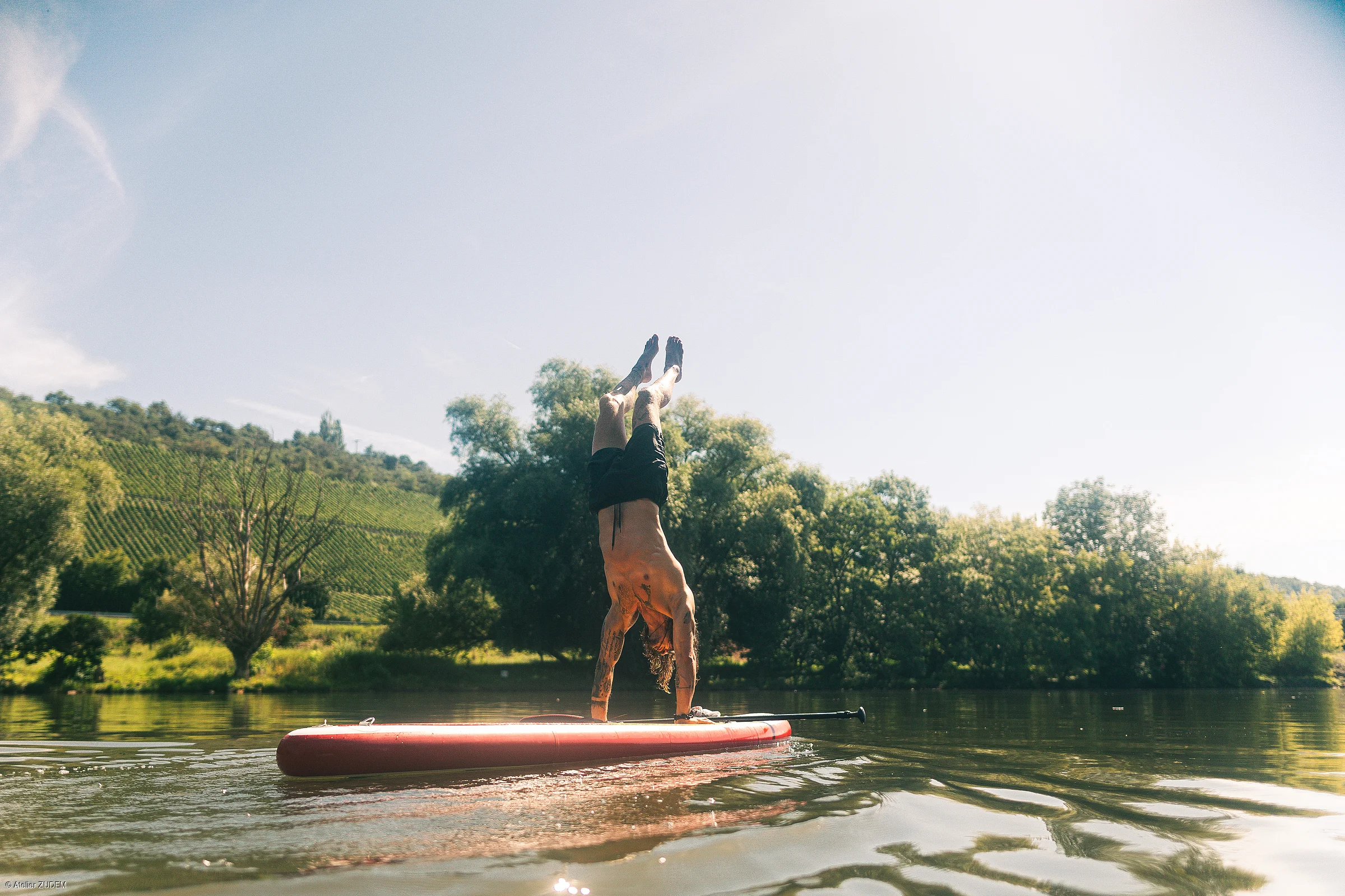 Mann macht Handstand auf einem roten Stand-up-Paddle-Board auf ruhigem Gewässer mit Bäumen im Hintergrund.