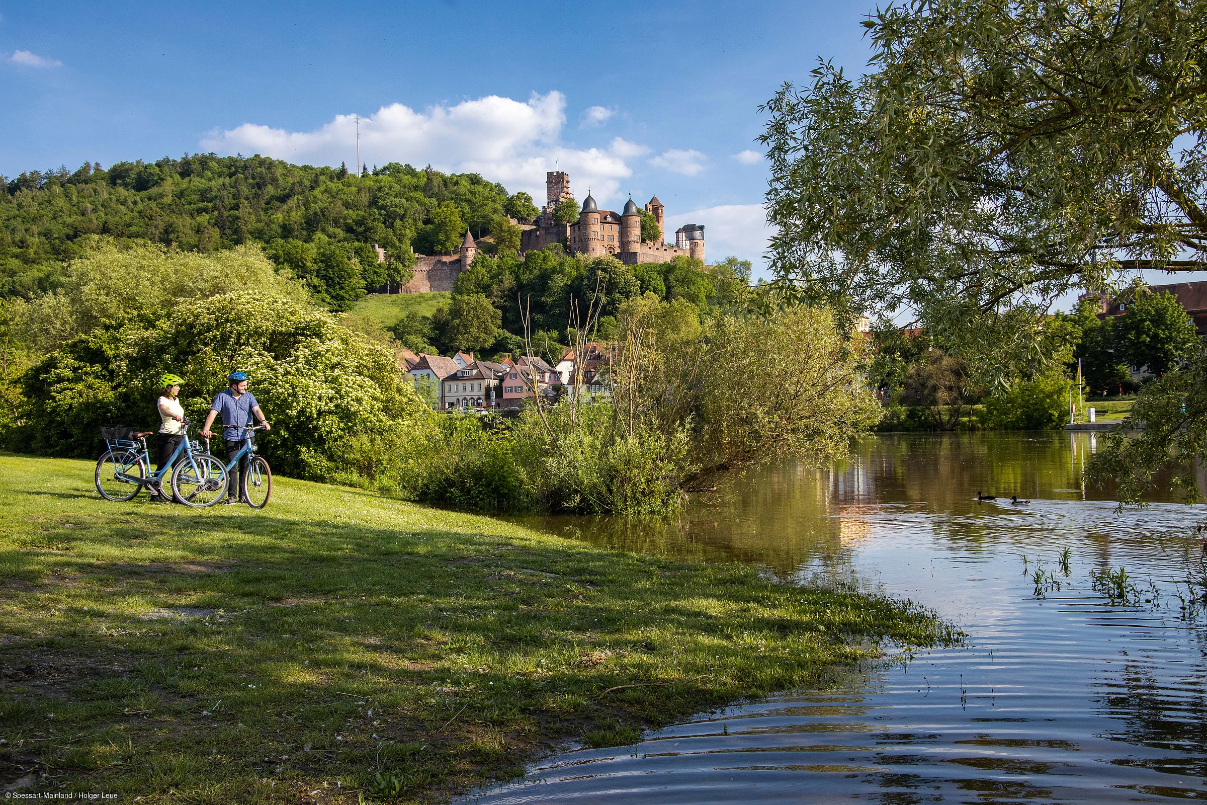 Zwei Personen mit Fahrrädern stehen auf einer Wiese neben einem Fluss, im Hintergrund ein bewaldeter Hügel mit einer Burg.