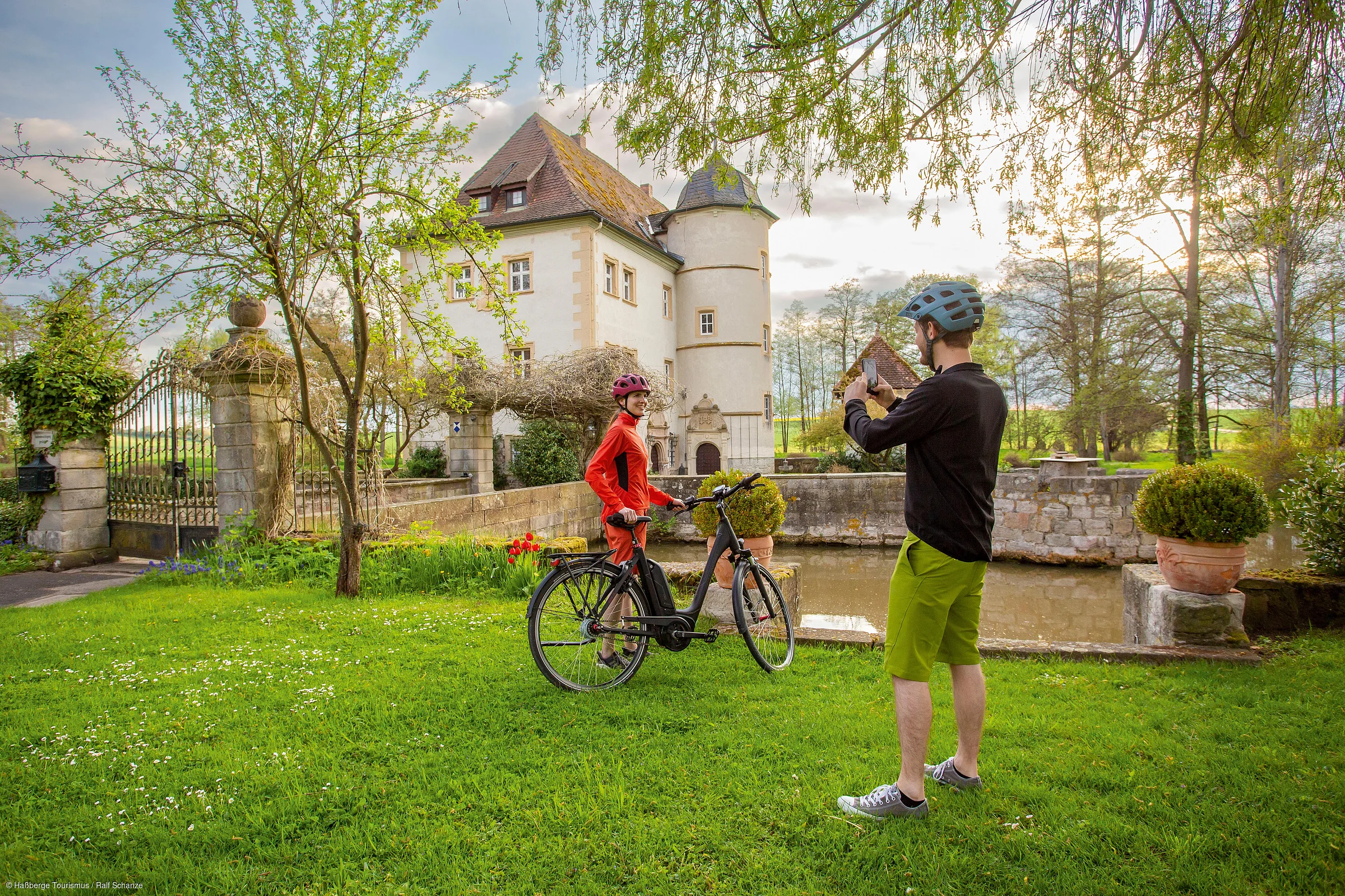 Zwei Personen mit Fahrradhelmen in einem Garten vor einem historischen Gebäude, eine macht ein Foto von der anderen.