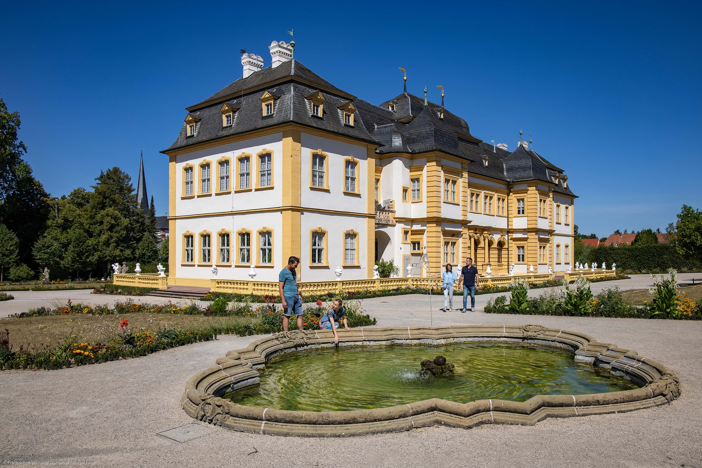 Barockschloss mit gelb-weißer Fassade, Brunnen im Vordergrund und vier Personen im Schlossgarten bei Sonnenschein