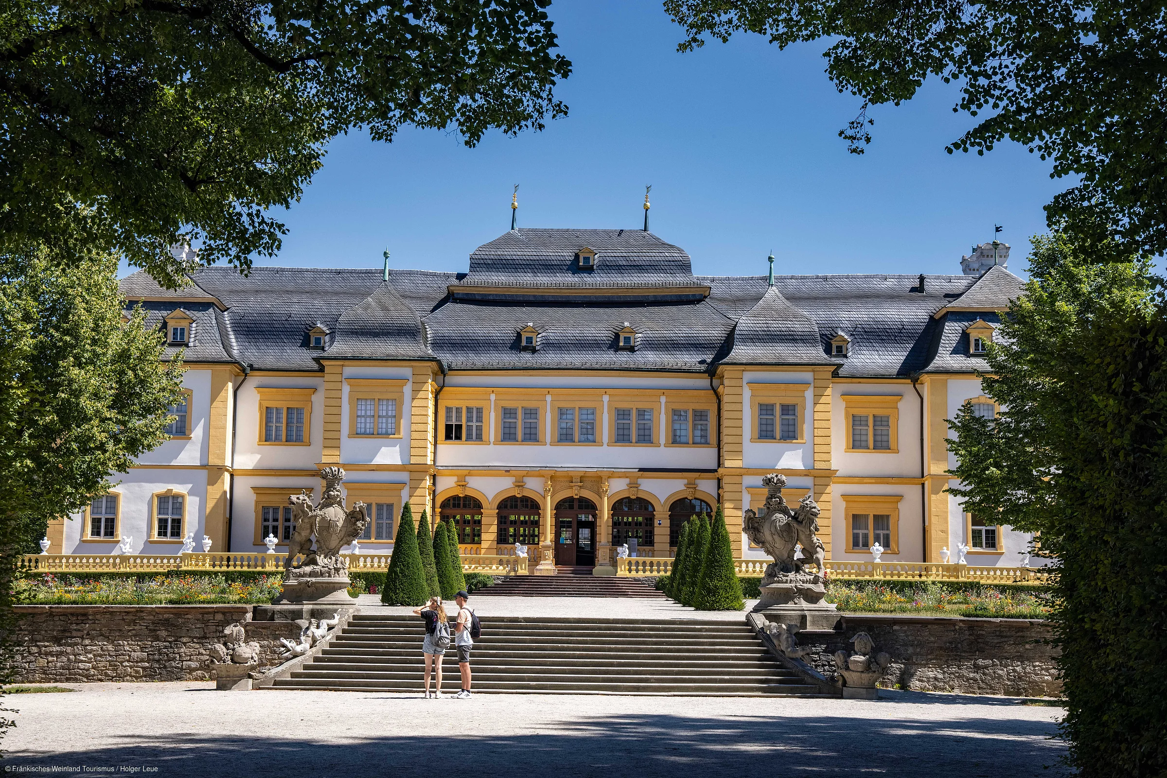 Barockschloss mit gelb-weißer Fassade, Treppe, Statuen und zwei Personen im Vordergrund bei blauem Himmel