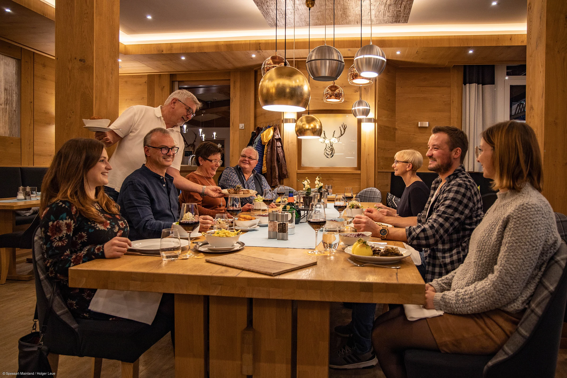 Acht Personen sitzen an einem Holztisch in einem Restaurant, ein Kellner serviert Essen.