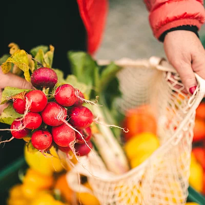 Person hält einen Bund Radieschen über eine Netztasche mit anderem Gemüse auf einem Markt.