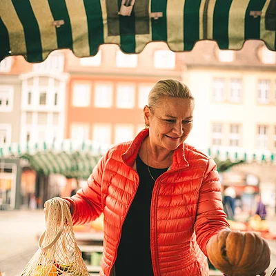 Frau in roter Jacke hält Kürbis auf Markt, trägt Netzbeutel mit Gemüse, Marktstände und Gebäude im Hintergrund.