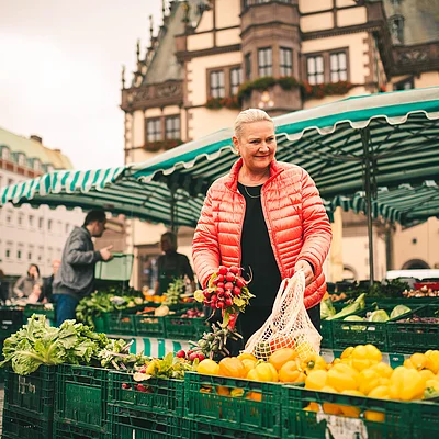 Frau mit roter Jacke kauft Radieschen auf einem Markt mit grün-weiß gestreiften Marktzeltplanen.