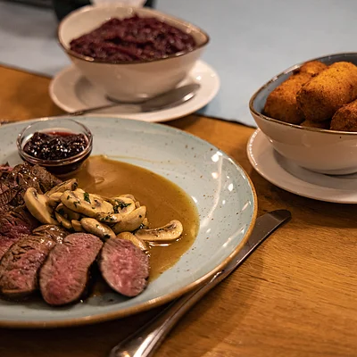 Teller mit gebratenem Fleisch, Pilzen und Sauce, daneben Schale mit Kroketten und Schale mit Rotkohl auf Holztisch