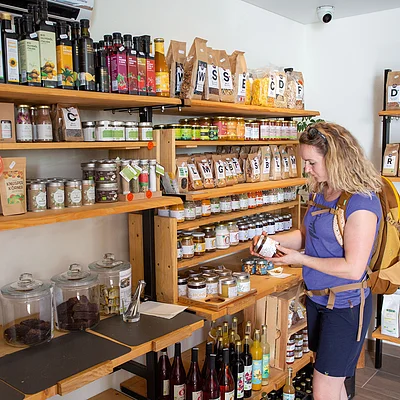 Frau mit Rucksack steht in kleinem Laden und betrachtet ein Glas mit Lebensmitteln vor Holzregalen voller Gläser und Packungen.
