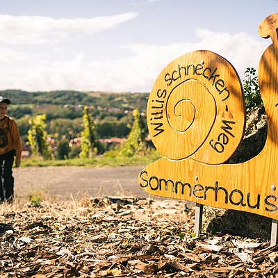 Holzschild in Schneckenform mit der Aufschrift „Willis Schnecken-Weg Sommerhausen“ neben einem Weg, im Hintergrund ein Junge und Landschaft.