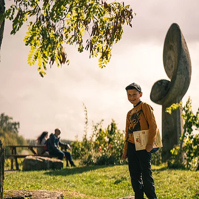 Junge mit Mütze steht auf Steinplatte im Park, hält Papier, im Hintergrund zwei sitzende Personen.