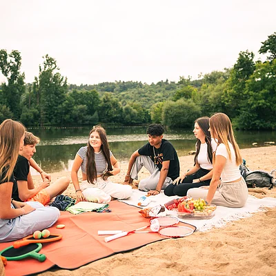 Sechs Jugendliche sitzen auf Decken am Sandstrand neben einem See mit Bäumen im Hintergrund.