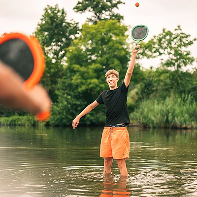 Zwei Jugendliche spielen mit Schlägern und Ball im flachen Wasser eines Sees, umgeben von Bäumen.
