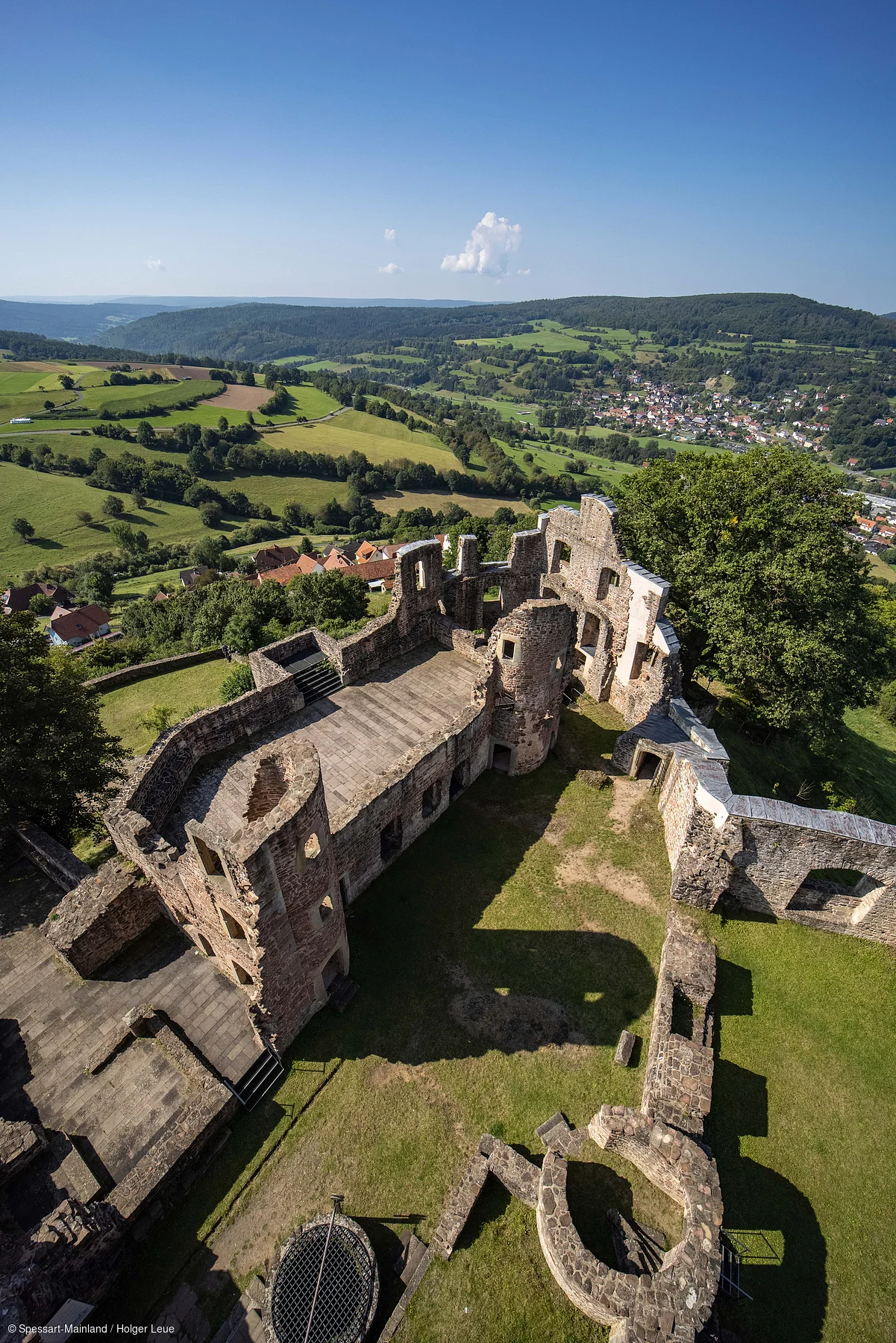 Blick von oben auf die Ruine einer Burg mit umliegender grüner Landschaft und Dorf im Hintergrund.