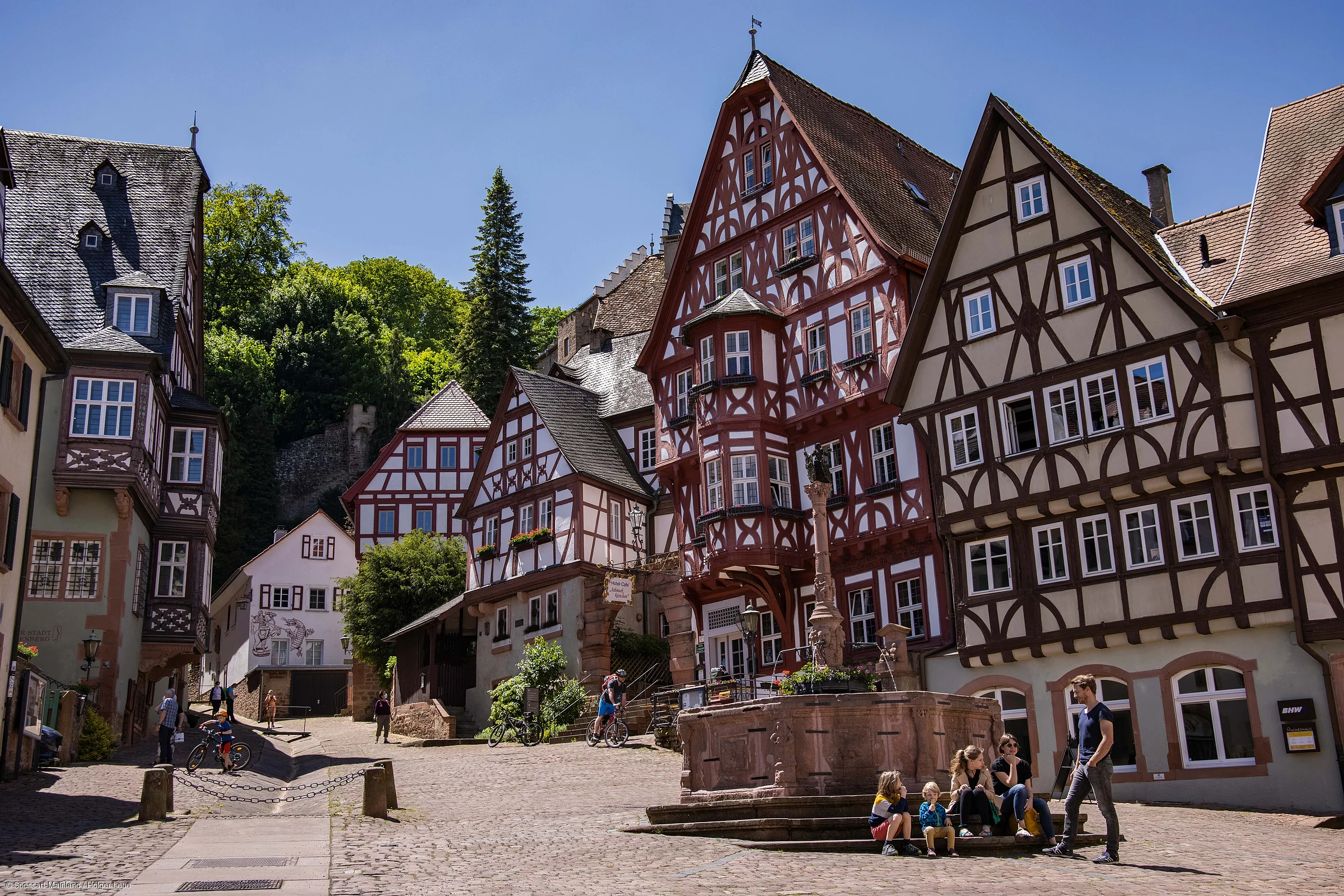 Historische Fachwerkhäuser an einem sonnigen Tag mit Menschen auf einem gepflasterten Platz und einem Brunnen.