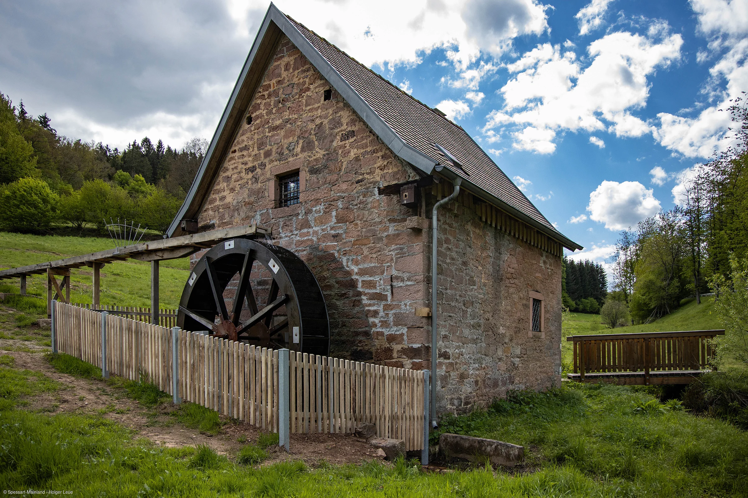 Altes Steinhaus mit Wasserrad und Holzzaun in grüner Landschaft unter bewölktem Himmel.