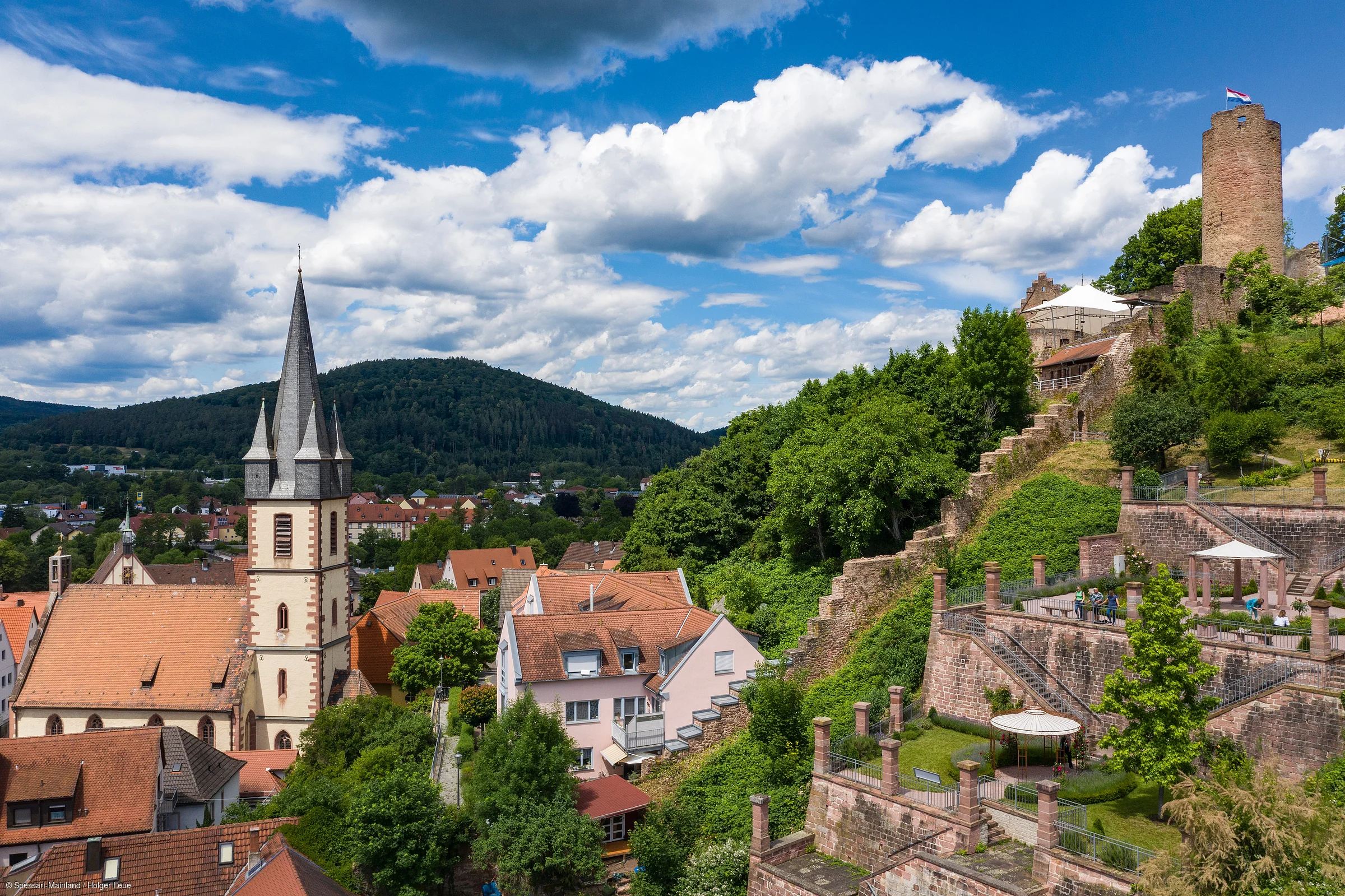 Blick auf eine Kirche mit spitzem Turm, Häuser, grüne Hügel und eine alte Burgmauer mit Turm und Flagge.