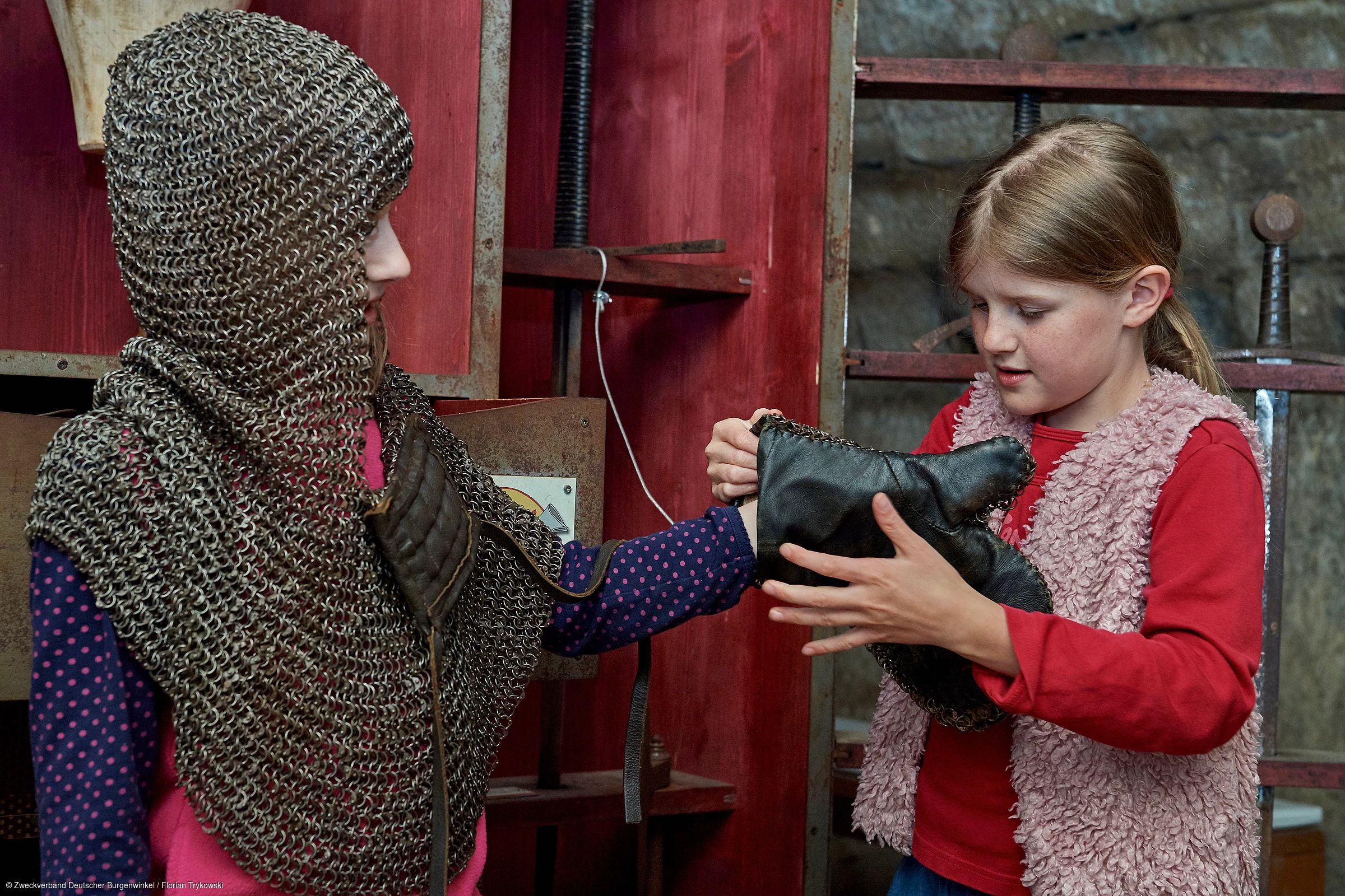 Zwei Mädchen, eines trägt Kettenhemd, das andere hilft beim Anziehen eines gepolsterten Handschuhs.