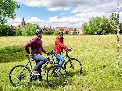 Zwei Personen mit Fahrradhelmen auf Mountainbikes auf einer Wiese vor einem Dorf mit Häusern und Kirche im Hintergrund.