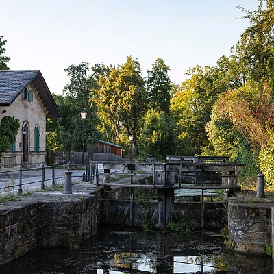 Altes Schleusentor mit Wasser, umgeben von Bäumen und einem Gebäude mit grünem Fensterladen.