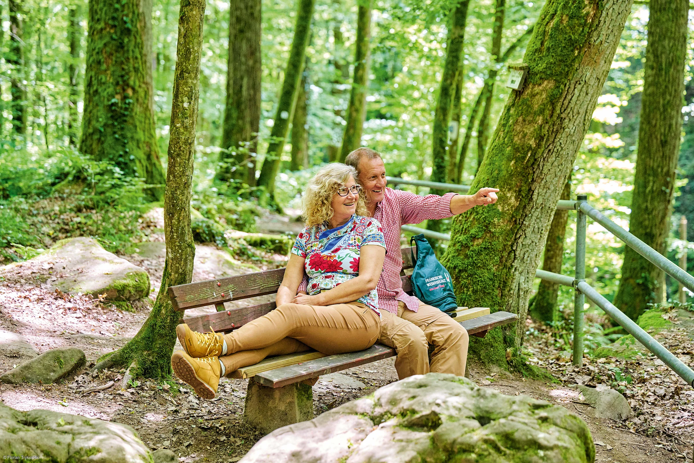 Älteres Paar sitzt auf einer Bank im Wald, Mann zeigt in die Ferne, Frau lehnt entspannt an ihm.