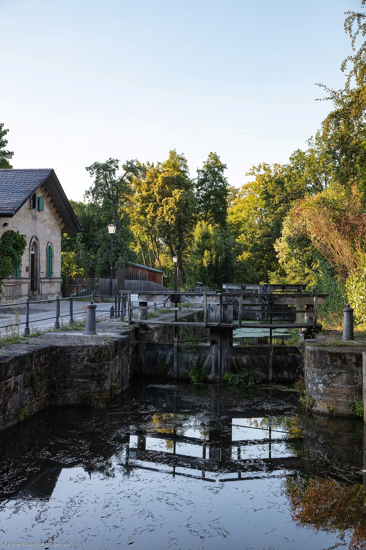 Altes Schleusentor mit Wasser, umgeben von Bäumen und einem Gebäude mit grünem Fensterladen.