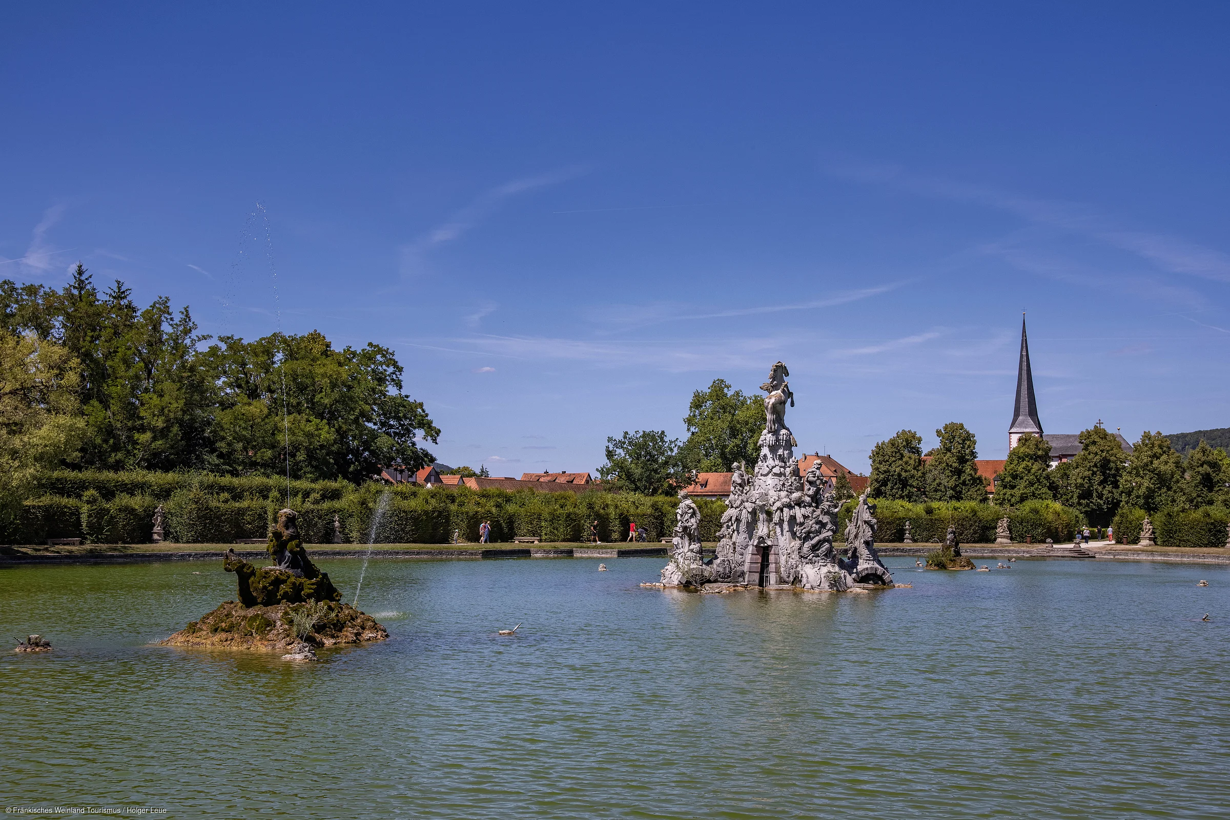 Wasserbecken mit mehreren Skulpturen, umgeben von Bäumen und einem Kirchturm im Hintergrund bei blauem Himmel.