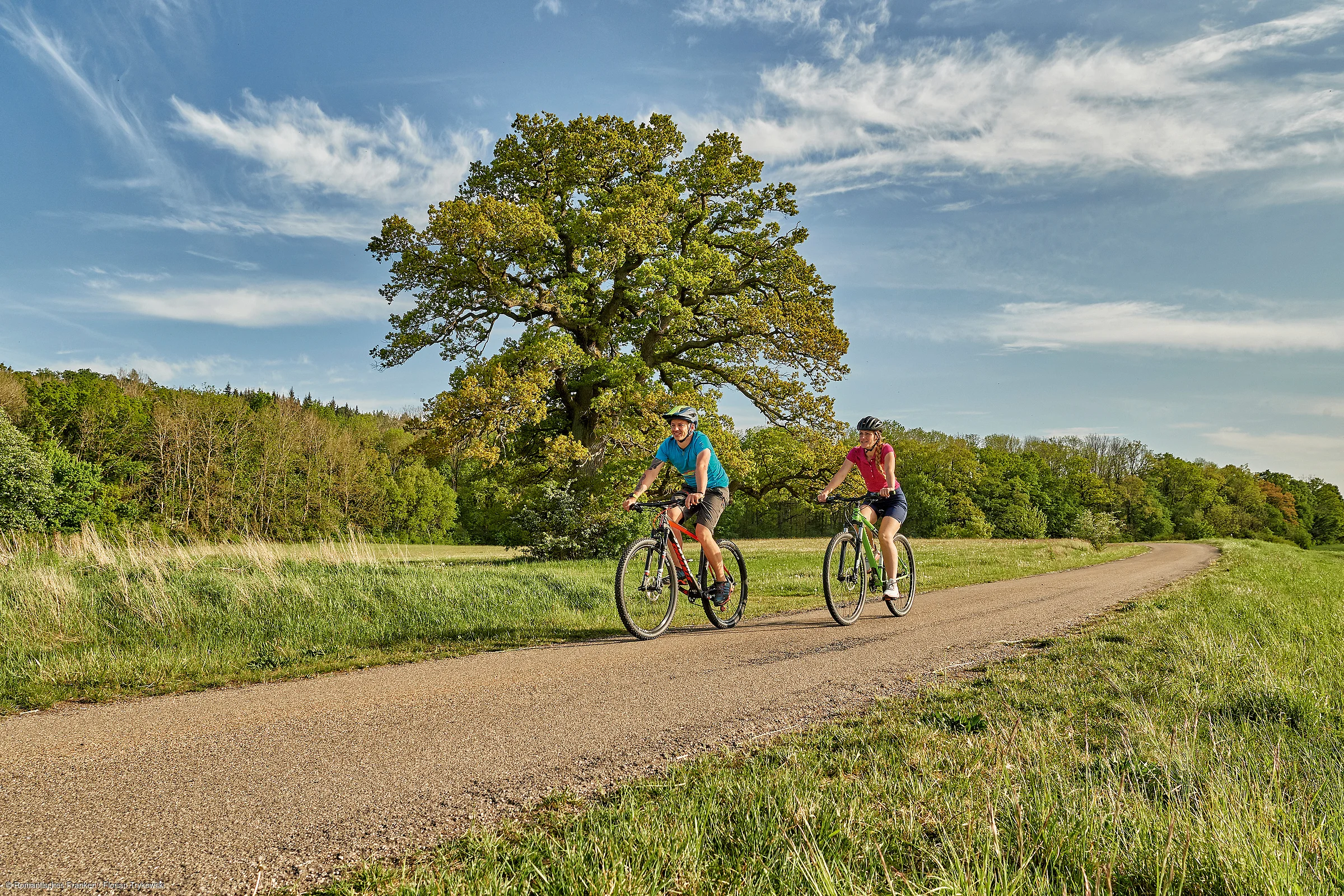 Zwei Personen fahren mit Fahrrädern auf einem Weg durch eine grüne Landschaft mit Bäumen und blauem Himmel.