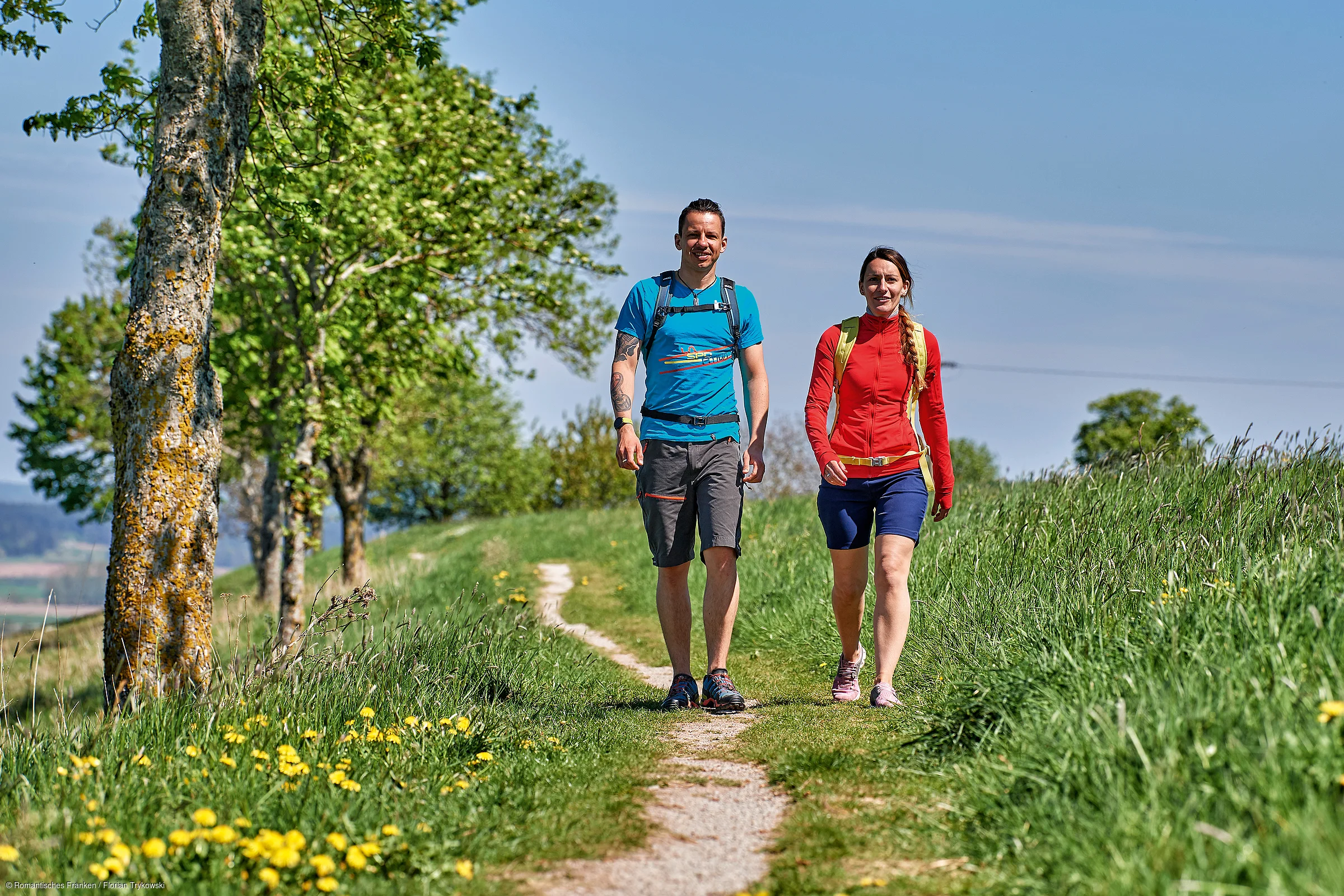 Zwei Personen wandern auf einem schmalen Weg zwischen grünen Wiesen und Bäumen bei klarem Himmel.
