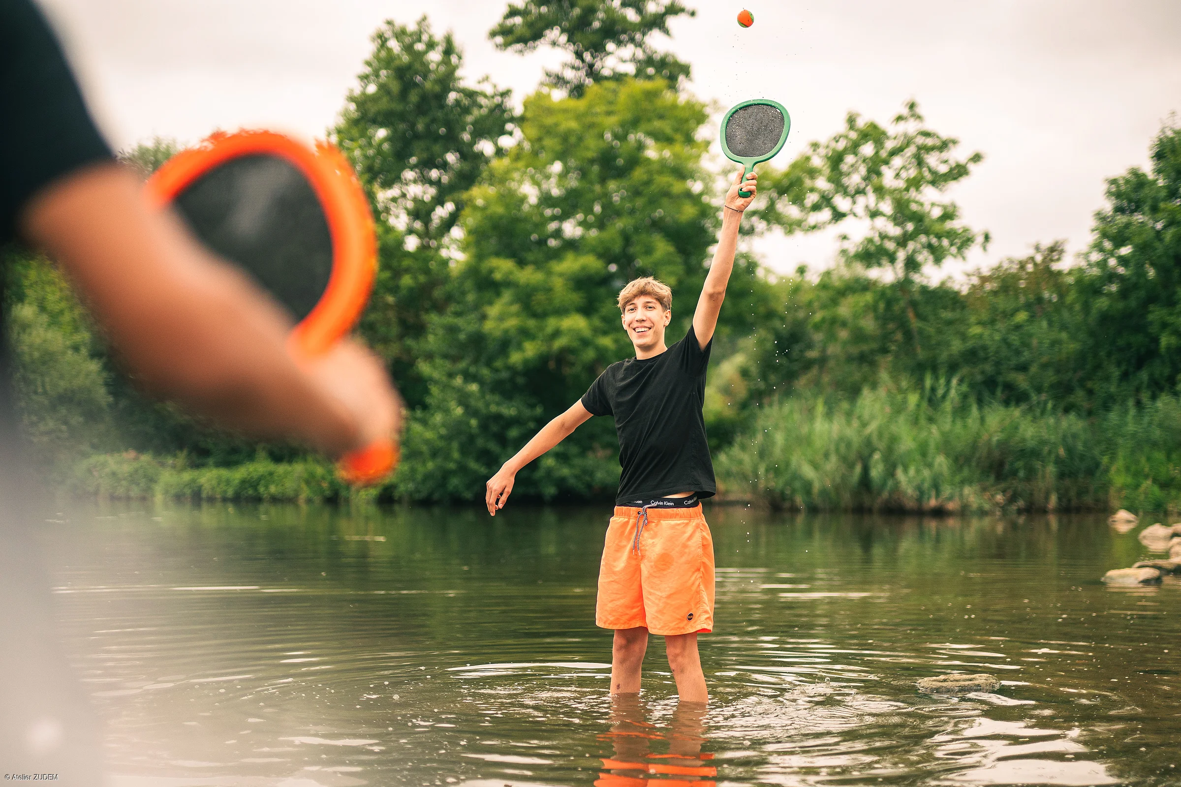 Zwei Jugendliche spielen mit Schlägern und Ball im flachen Wasser eines Sees, umgeben von Bäumen.