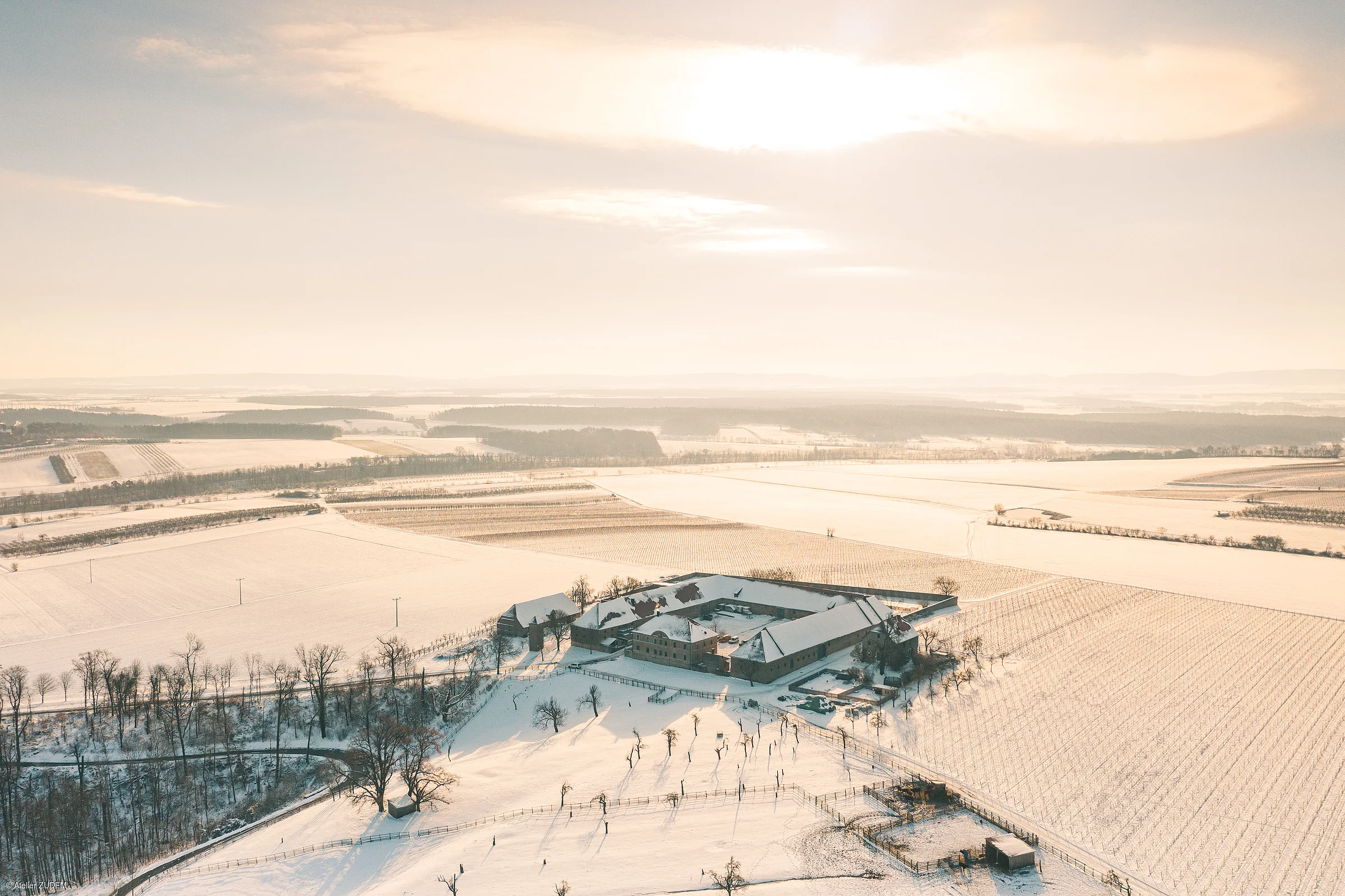 Luftaufnahme eines verschneiten Bauernhofs mit Feldern und Bäumen bei Sonnenlicht im Winter.