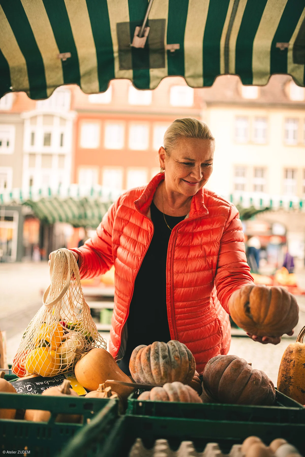 Frau in roter Jacke hält Kürbis auf Markt, trägt Netzbeutel mit Gemüse, Marktstände und Gebäude im Hintergrund.